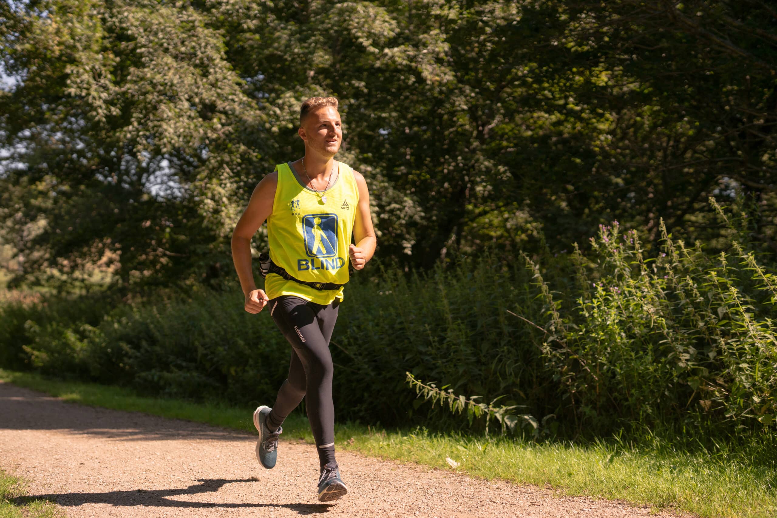 Edis running along a sunlit path surrounded by greenery, wearing a bright yellow vest with the word 'BLIND' and an icon symbolizing blindness. He is smiling, focused, and appears confident as he enjoys the run.