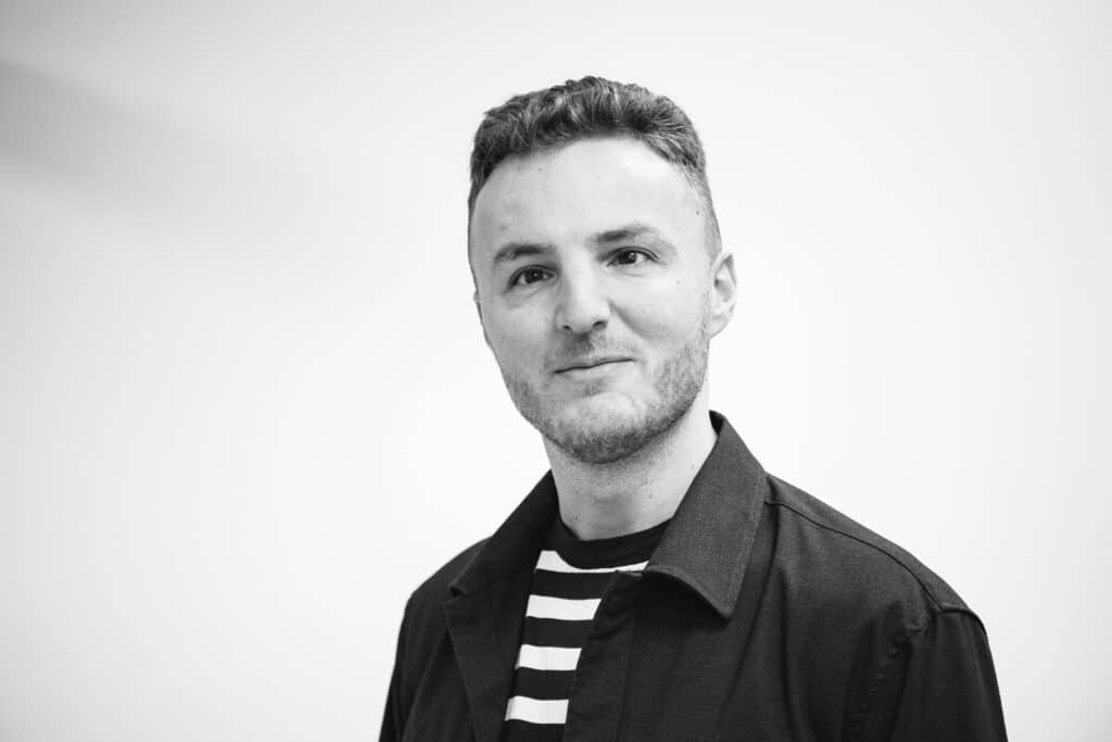 A black and white portrait of Edis, who has short, curly hair and a trimmed beard. He is wearing a striped shirt under a dark jacket, looking slightly off-camera with a subtle smile. The background is plain and light-colored.