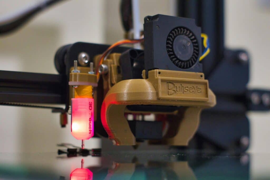 Close-up of a 3D printer's extruder assembly featuring a red-lit 3D Touch auto-bed leveling sensor attached to the nozzle. Above the nozzle is a cooling fan housed in a black casing, mounted on a 3D printed bracket labeled 'Bullseye.' The components are positioned above a smooth glass print bed, with the sensor's reflection visible on the bed's surface.