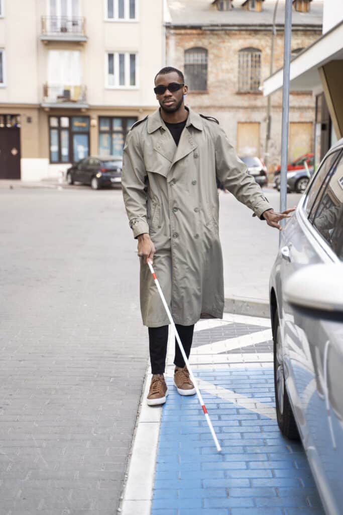 A man wearing a beige trench coat and dark sunglasses walks in an urban area, using a white cane for navigation and touching a parked car for guidance.