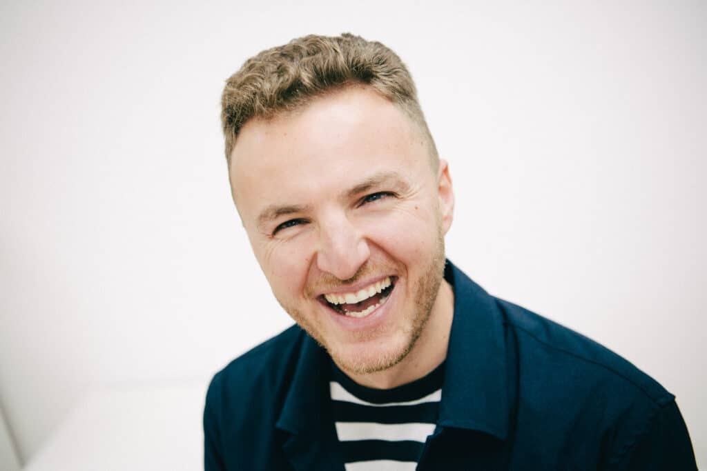 Edis smiling broadly, wearing a dark jacket over a striped shirt. He has short, curly hair and a trimmed beard, with a bright and cheerful expression on his face. The background is plain and light-colored.