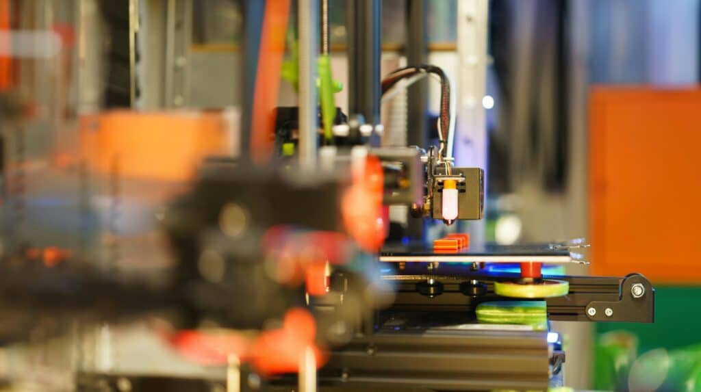 Side view of a multi-extruder 3D printer in operation, with several extruders aligned along a rail. The closest extruder is out of focus, while the farther one is in sharp focus, featuring a glowing component. The print bed below holds a partially printed object, with various mechanical components and colorful elements visible in the background.