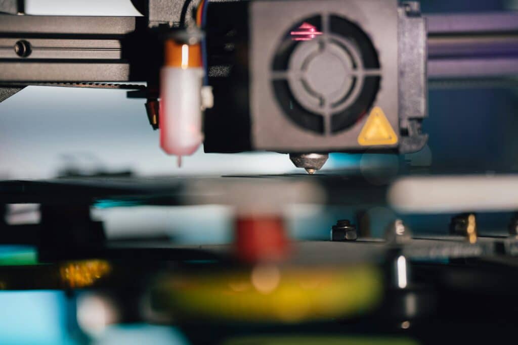 Close-up of a 3D printer extruder nozzle positioned above the print bed, with surrounding mechanical components, including a cooling fan and wiring, in soft, neutral lighting.