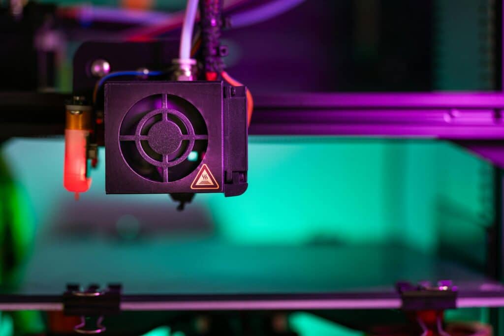 Close-up of a 3D printer extruder with a fan and a hot surface warning label, illuminated by vibrant purple and green lighting, with the print bed blurred in the background.