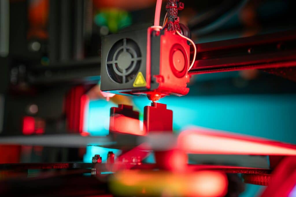 Close-up of a 3D printer extruder nozzle depositing filament, surrounded by mechanical components and illuminated by red and turquoise lighting.